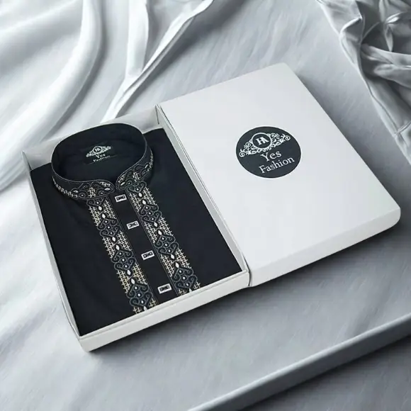 A premium black Cotton fabric men's panjabi neatly arranged inside a white gift box, featuring delicate golden and white embroidered design on the collar and both sides of the front placket with golden rectangular metal buttons; the box lid displays a black circular logo with the brand name "YES Fashion", also visible on the label inside the collar, photographed on a white fabric background.